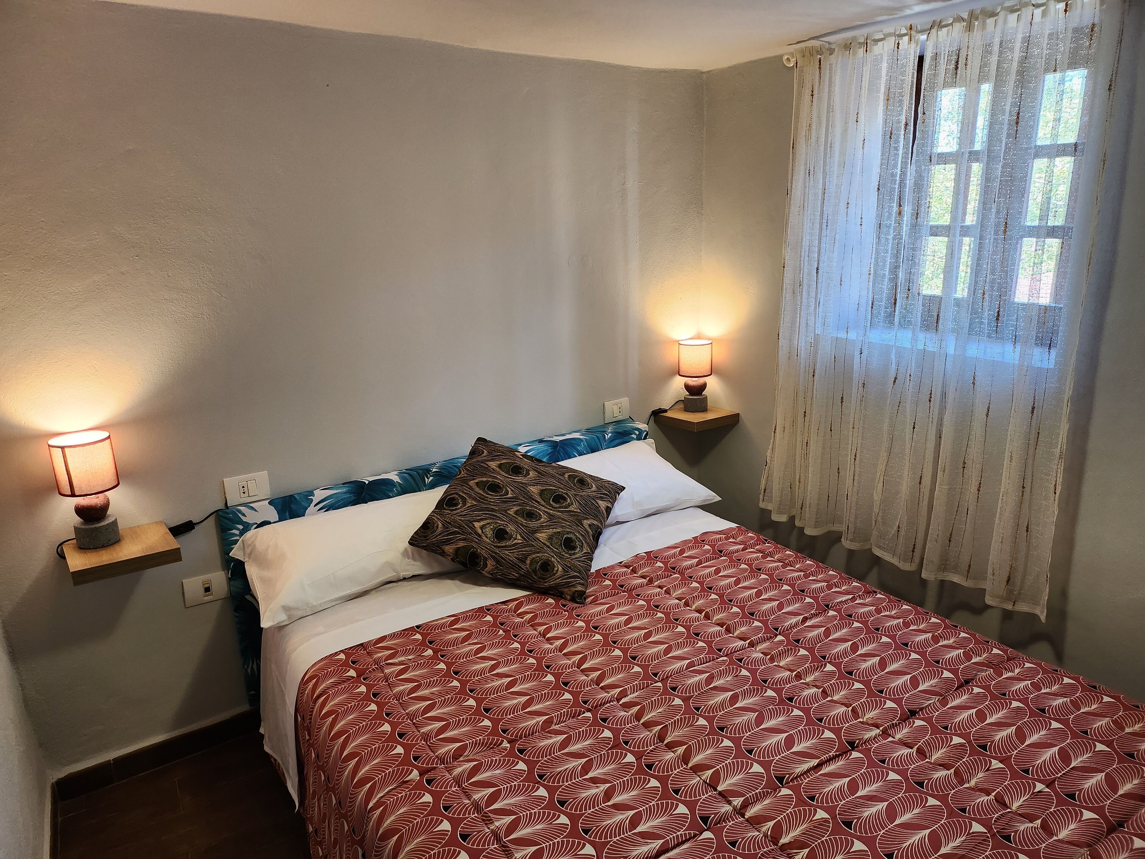 A bedroom with a bed, two bedside lamps, a window with sheer curtains, and a wall-mounted shelf.