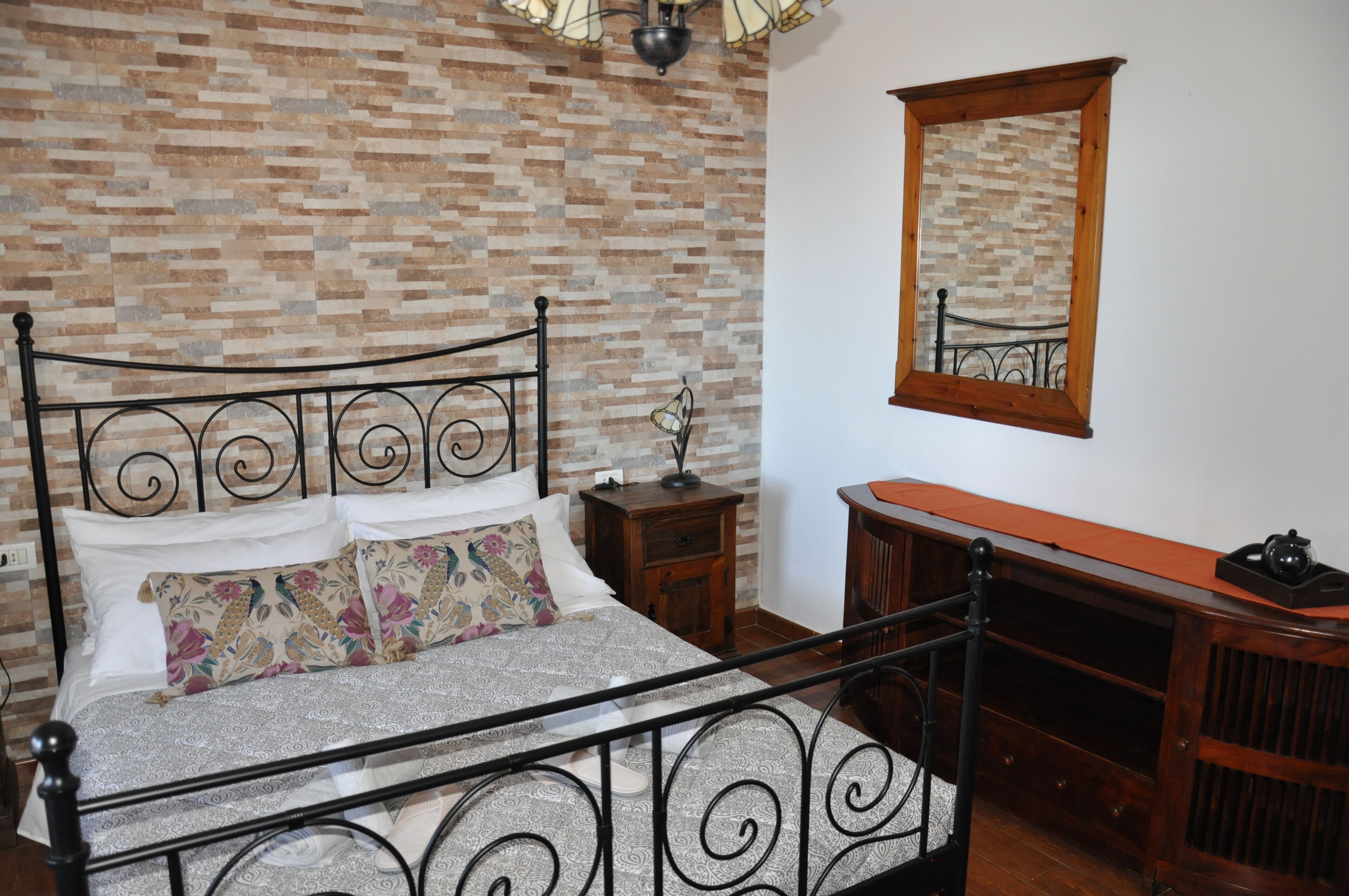 Un dormitorio con pared de ladrillo, cama con estructura metálica, mesita de noche de madera, cómoda y espejo.