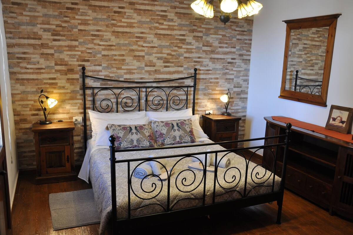 Een slaapkamer met een metselwerk muur, een metalen bedframe, twee nachtkastjes met lampen, een houten dressoir en een spiegel aan de muur.