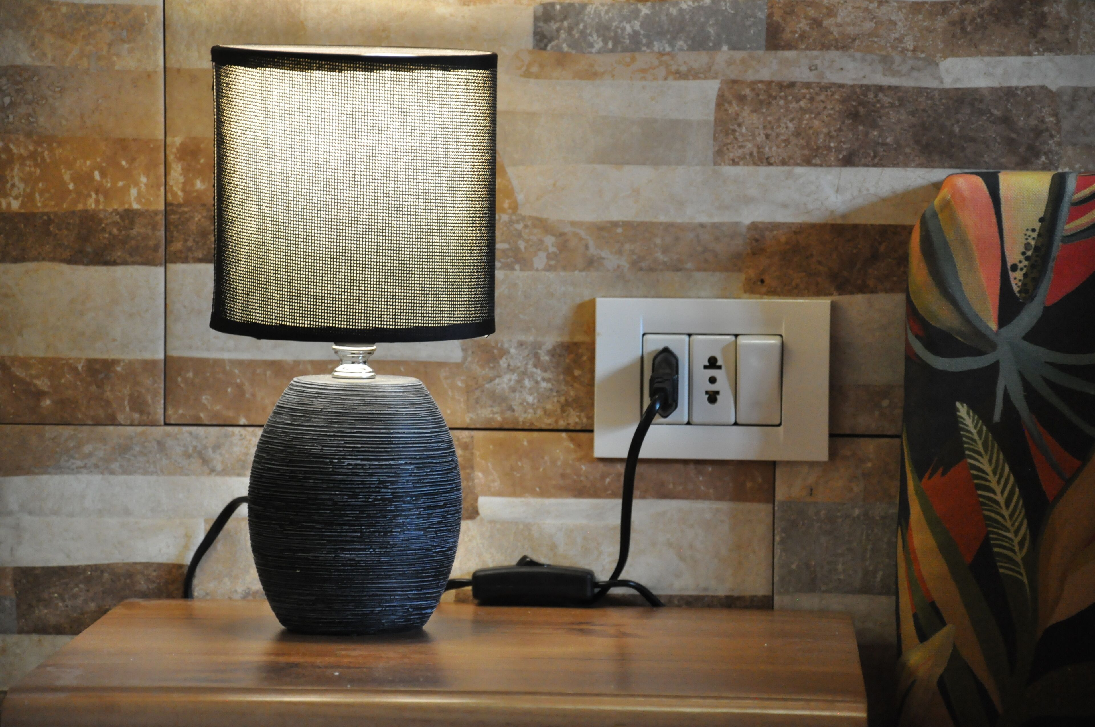En lampe med en struktureret skærm på et træbord, en stikkontakt, og en farverig stol.