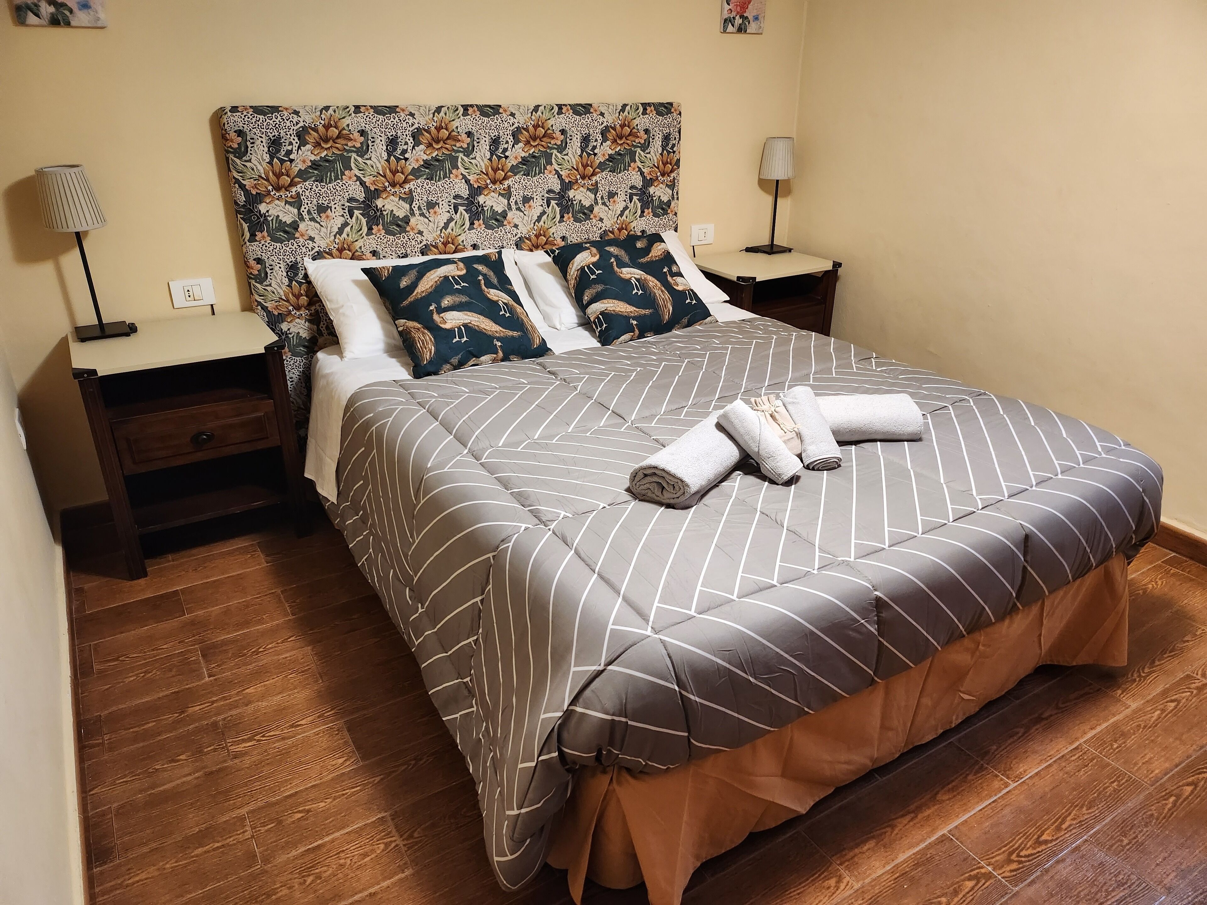 A neatly made bed with striped bedding and rolled towels, a floral-patterned headboard, and wooden flooring.