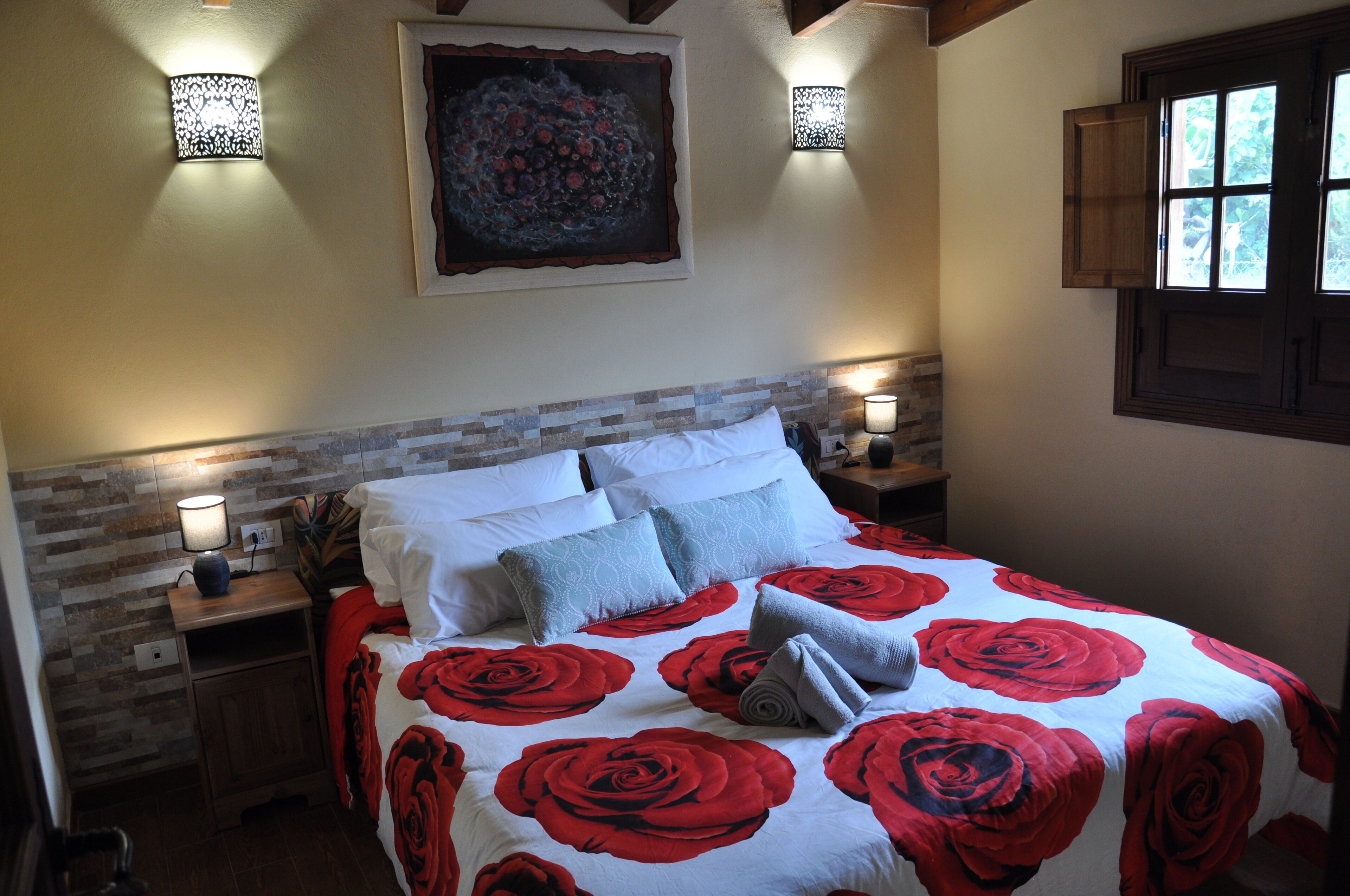 Una cama bien tendida con una colcha floral roja y blanca, dos lámparas de noche, una mesita de noche y una obra de arte colgada en la pared.