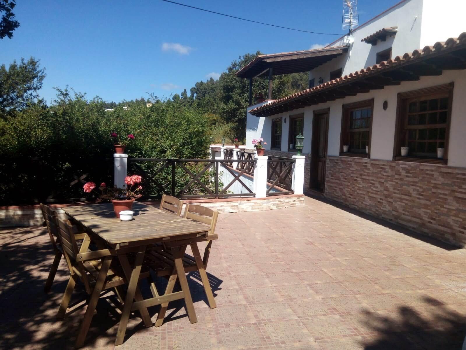 An outdoor patio with a wooden table and chairs, surrounded by potted plants and a stone wall.