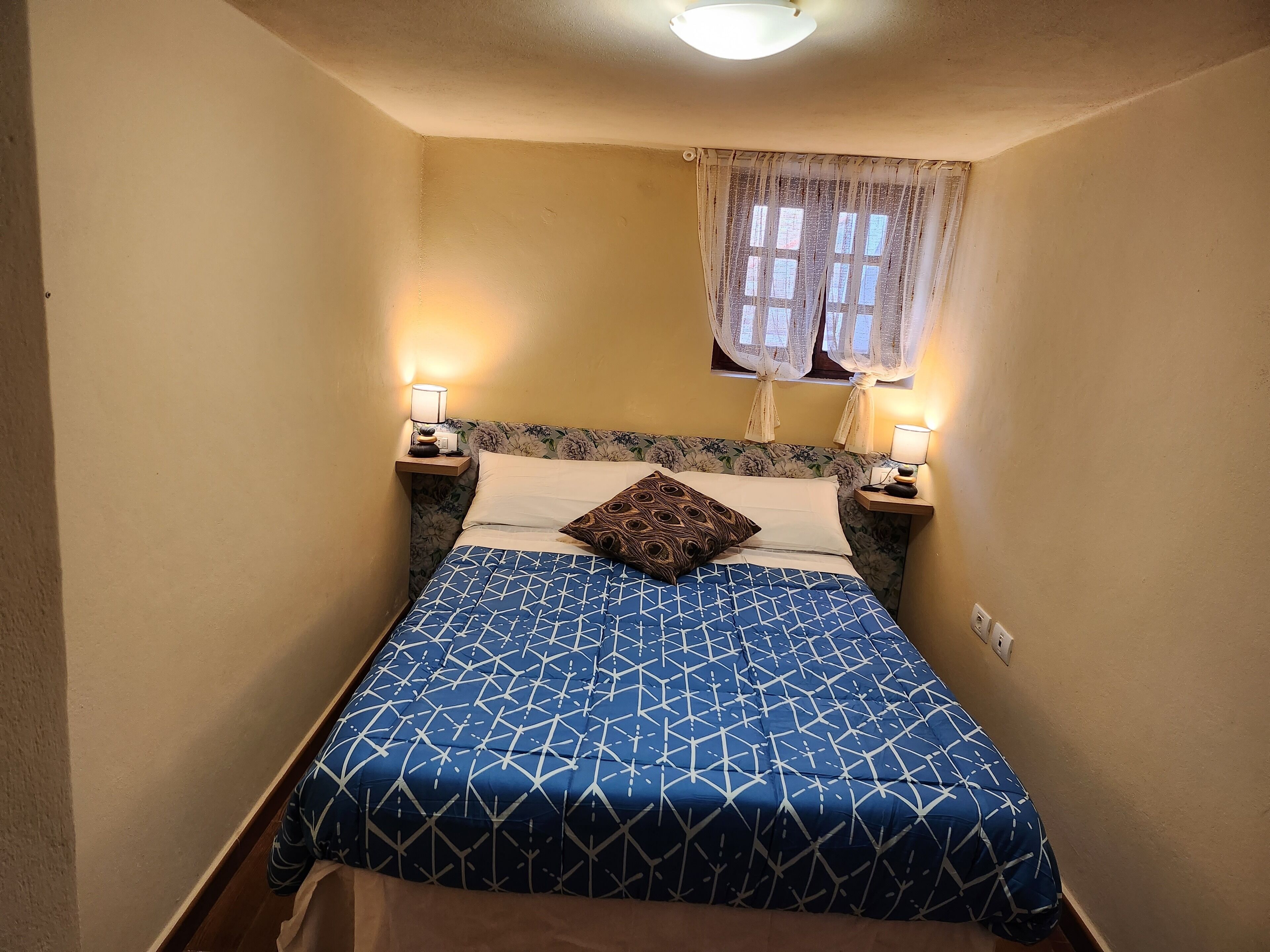 A narrow room with a bed, two bedside tables, and a window with curtains.