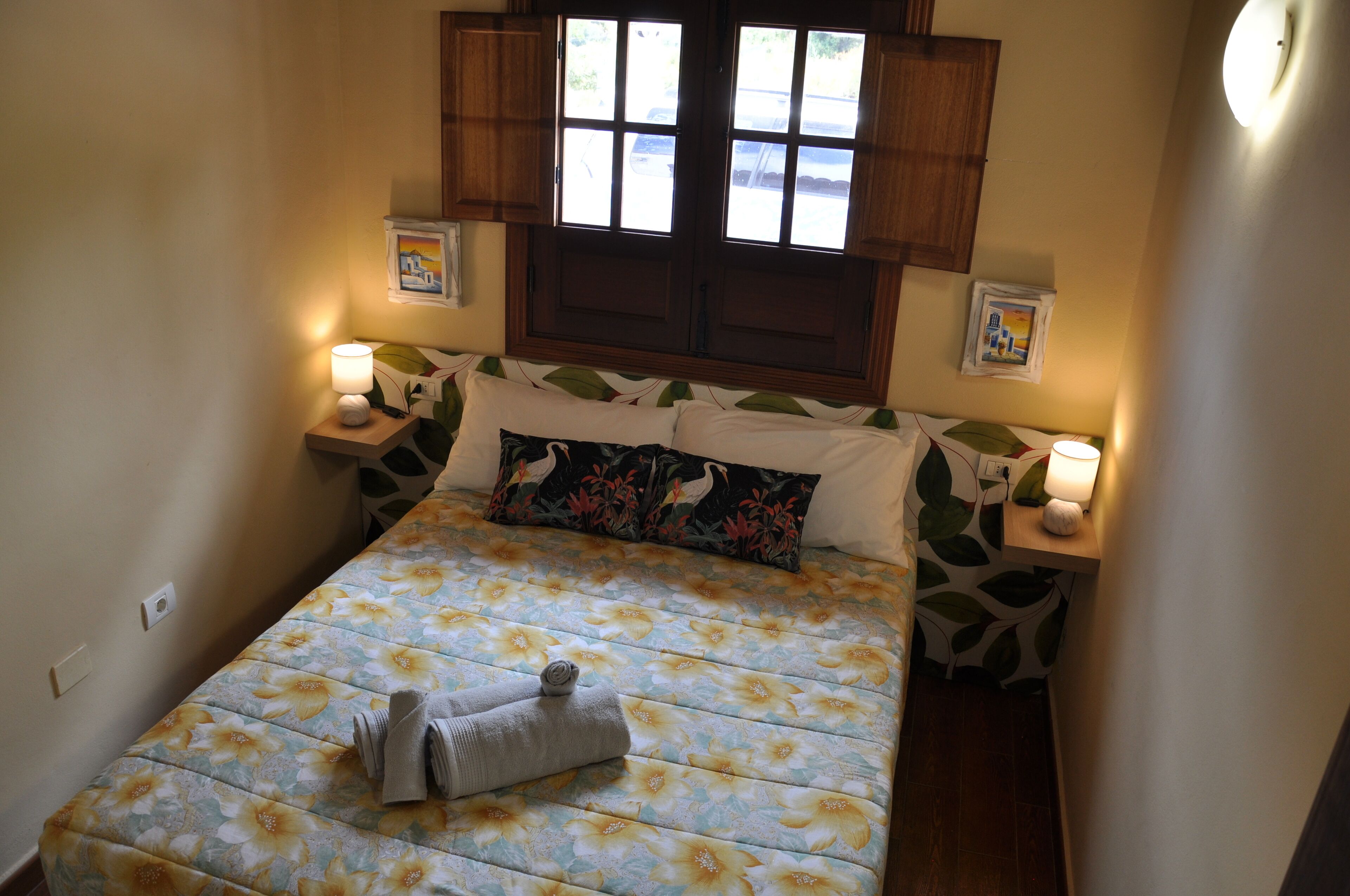 Une petite chambre à coucher accueillante, avec une couvre-lit à motifs floraux, un lit bien fait sur lequel est posée une serviette enroulée, des armoires en bois au-dessus du lit et deux lampes de chevet.