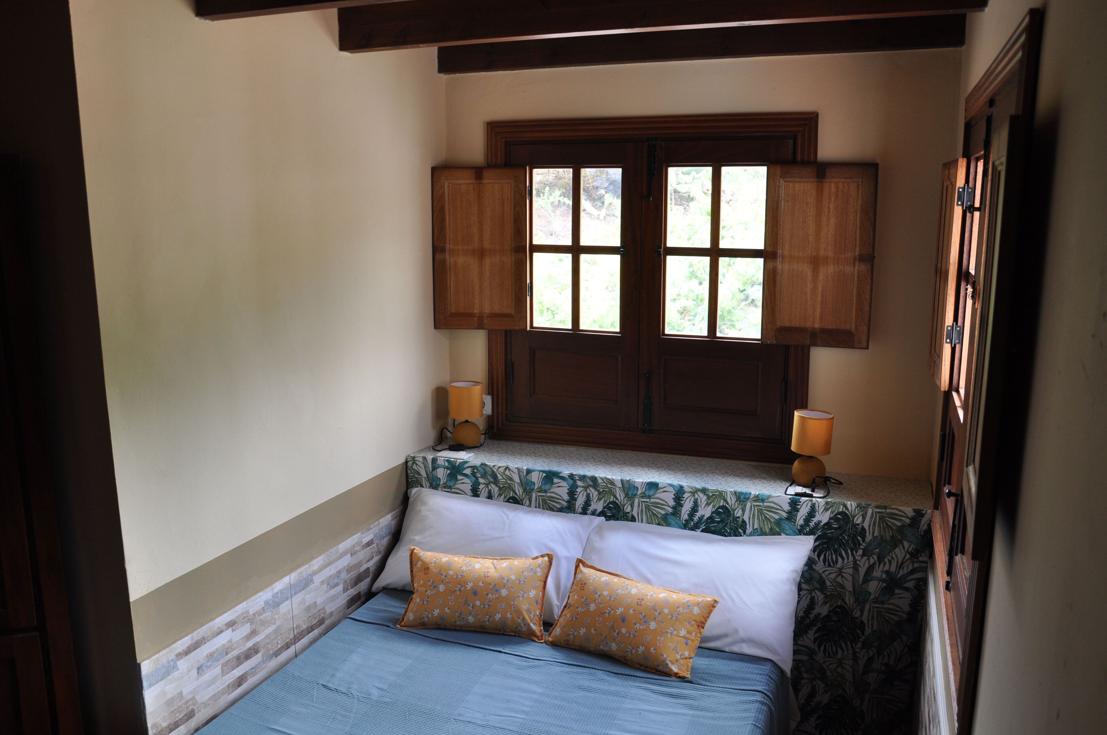 Un dormitorio con una cama, dos mesitas de noche, un ventanal con contraventanas de madera y un armario empotrado.