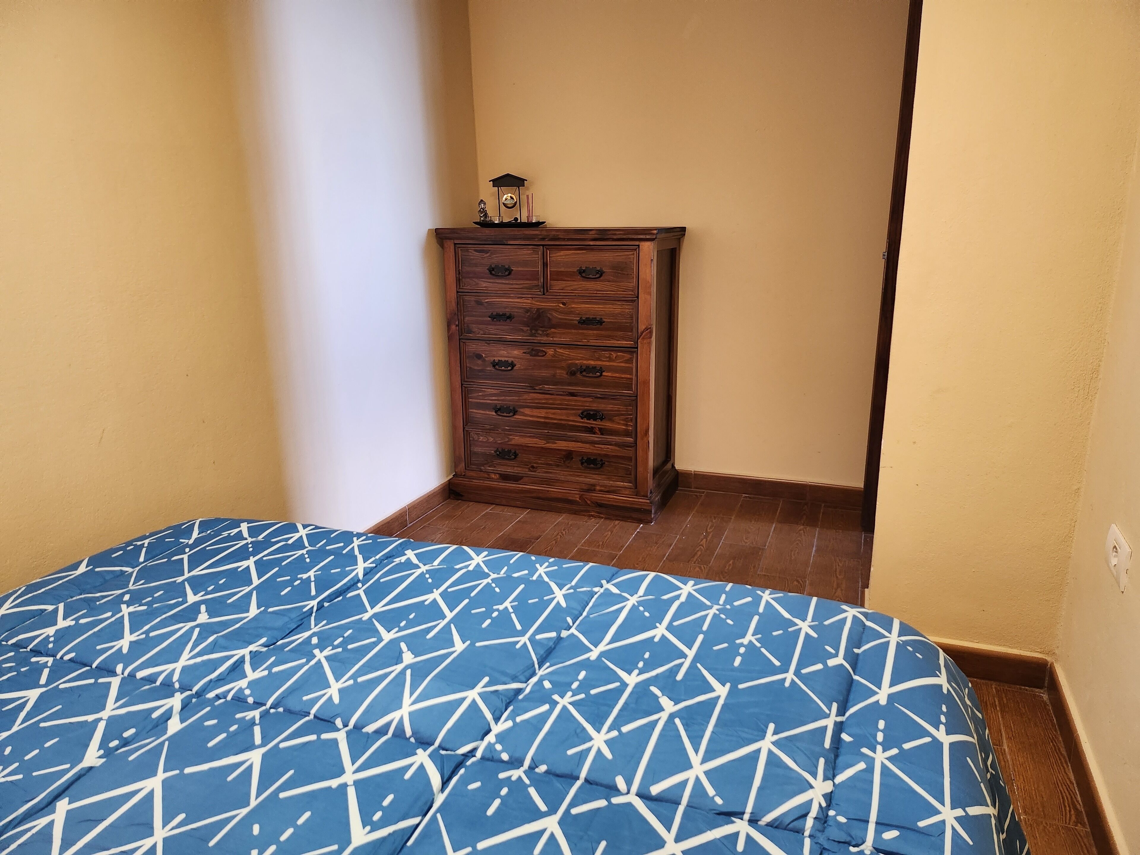 Een slaapkamer met een houten dressoir, een bed met een blauw bedsprei met patroon en een effen muur.