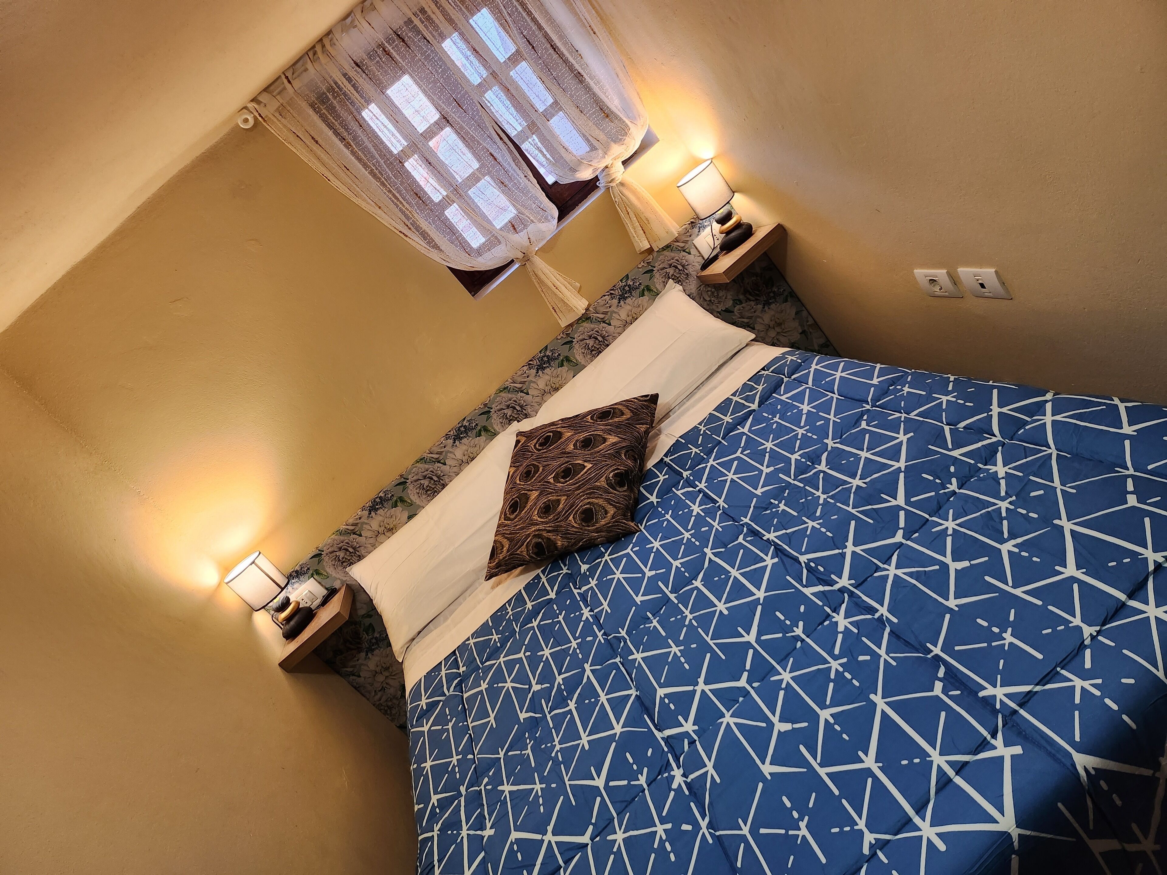 Una cama con un edredón azul estampado, un cojín decorativo y dos lámparas de noche.