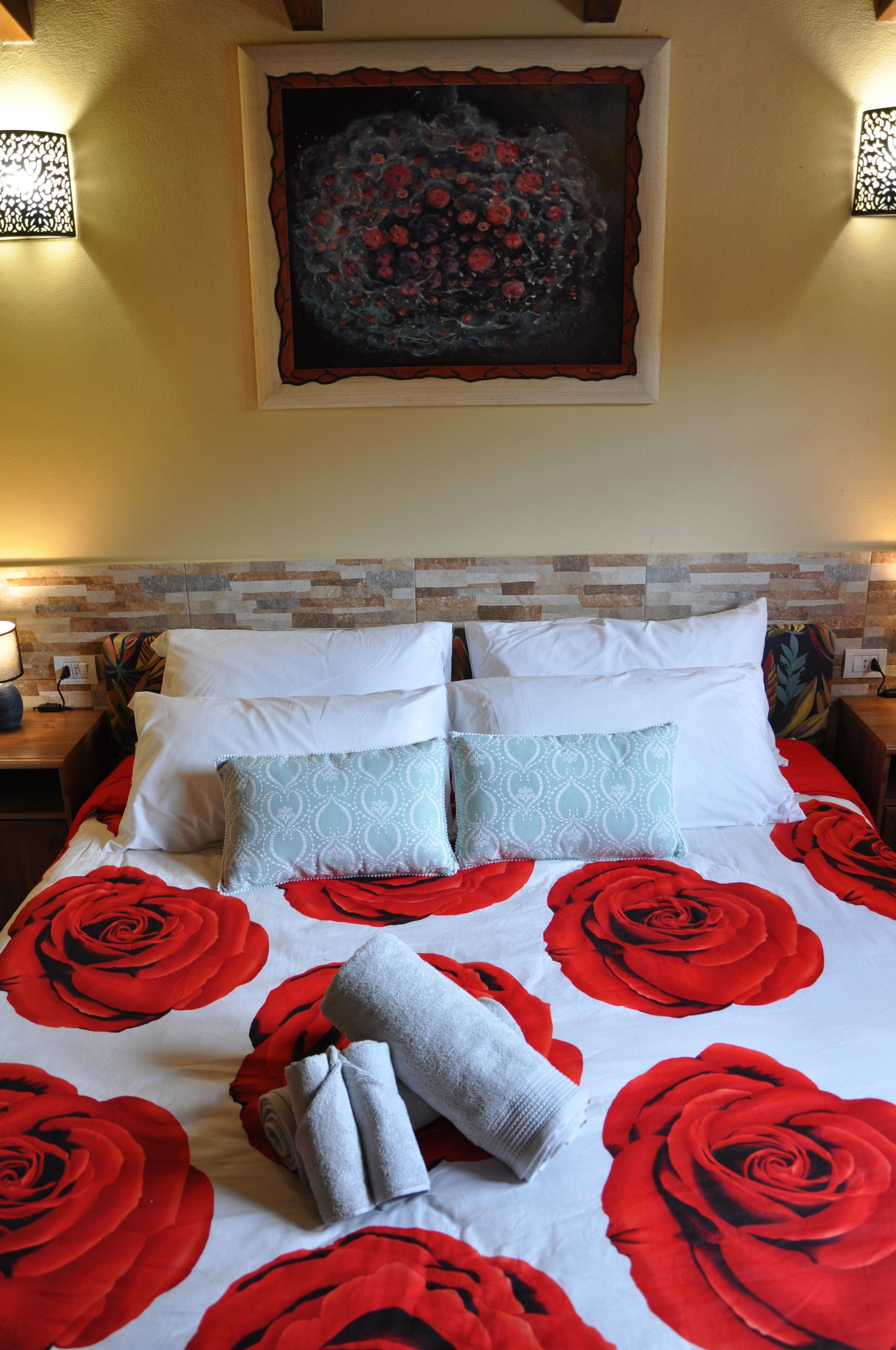 A hotel room with a bed featuring a red and white floral bedspread, two white pillows, and a decorative painting above the bed.