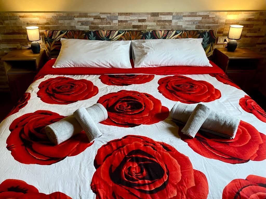 A hotel room with a bed featuring a red rose pattern bedspread, two white pillows, and two rolled towels on the bed.