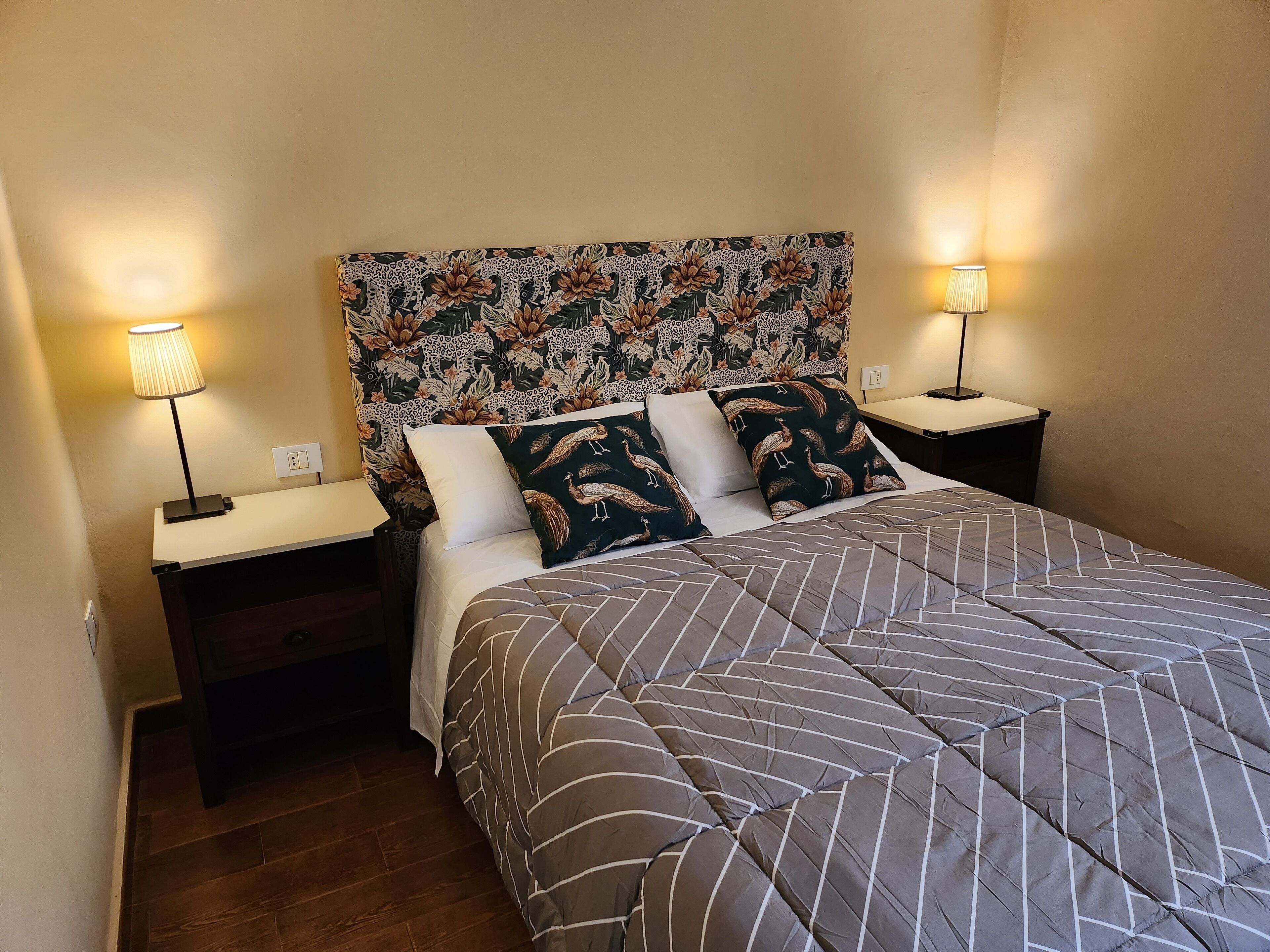 A neatly made bed with striped bedding, two pillows, and a floral-patterned headboard. There are two bedside tables with lamps on either side of the bed.