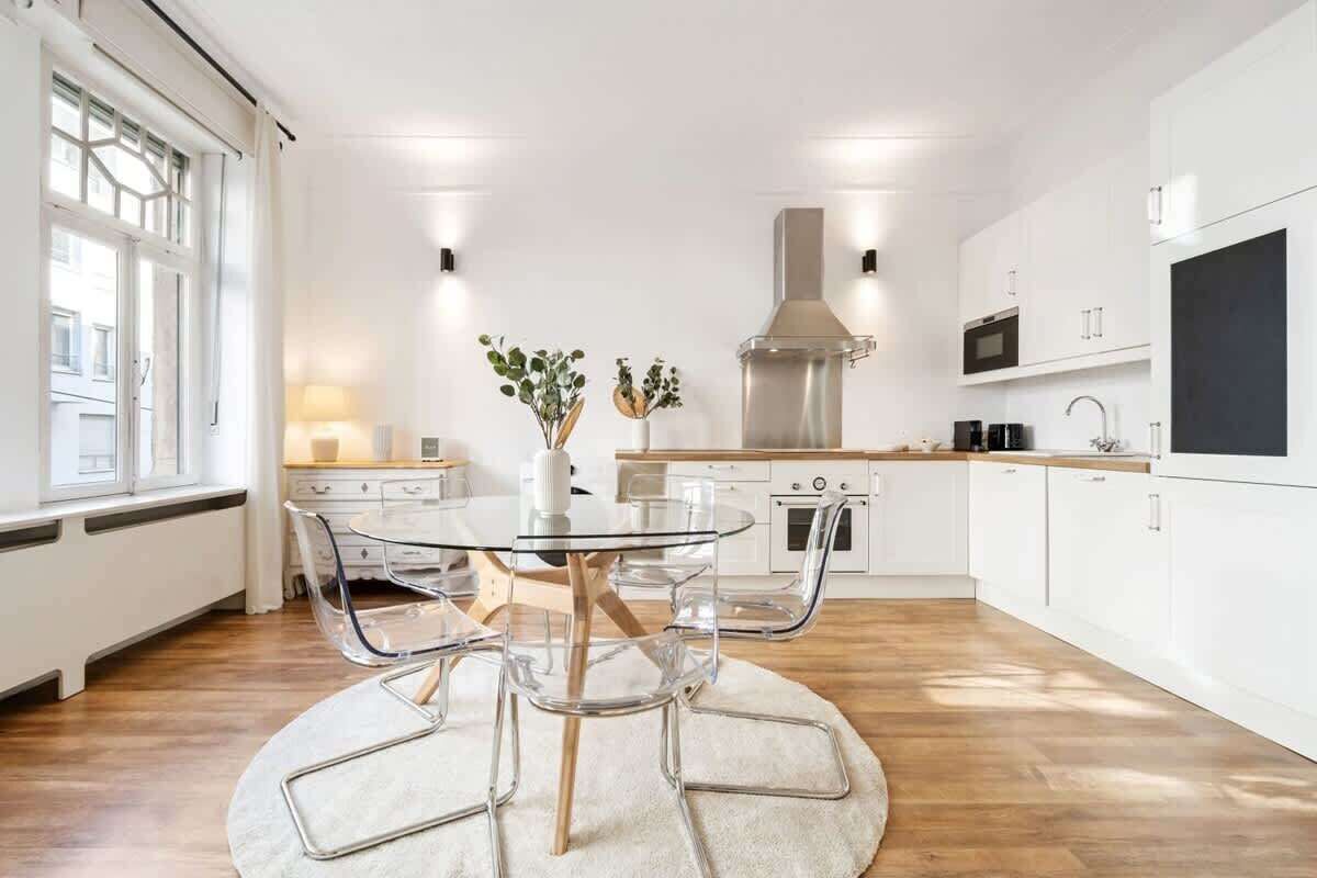 Une cuisine moderne avec des meubles blancs, une hotte en acier inoxydable et une table ronde en verre, accompagnée de chaises.