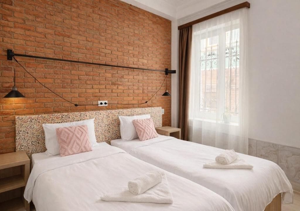 Una habitación de hotel con dos camas, una pared de ladrillo, una ventana con cortinas y una lámpara colgante.