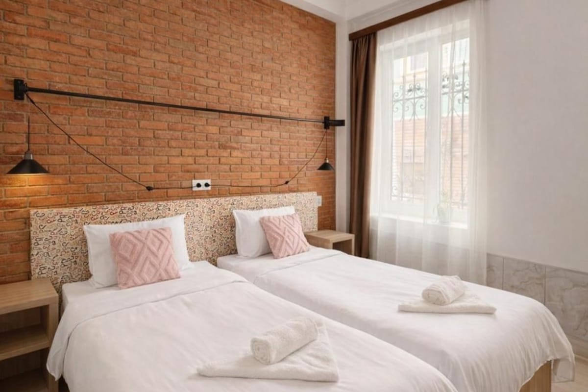 A hotel room with two beds, a brick wall, a window with curtains, and a hanging lamp.
