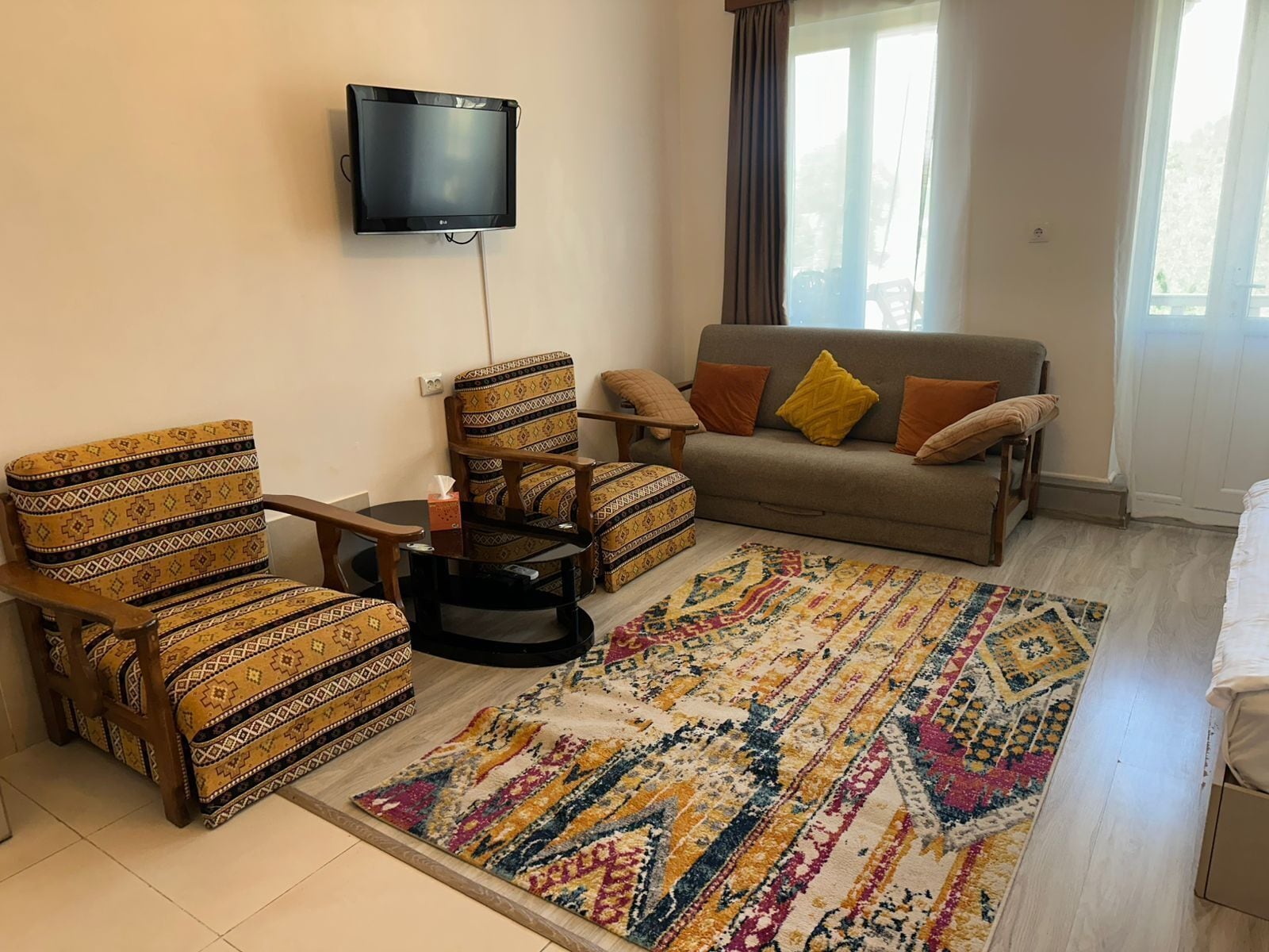 Una sala de estar con un sofá, dos sillones estampados, una mesa de centro, un televisor montado en la pared y una alfombra colorida.