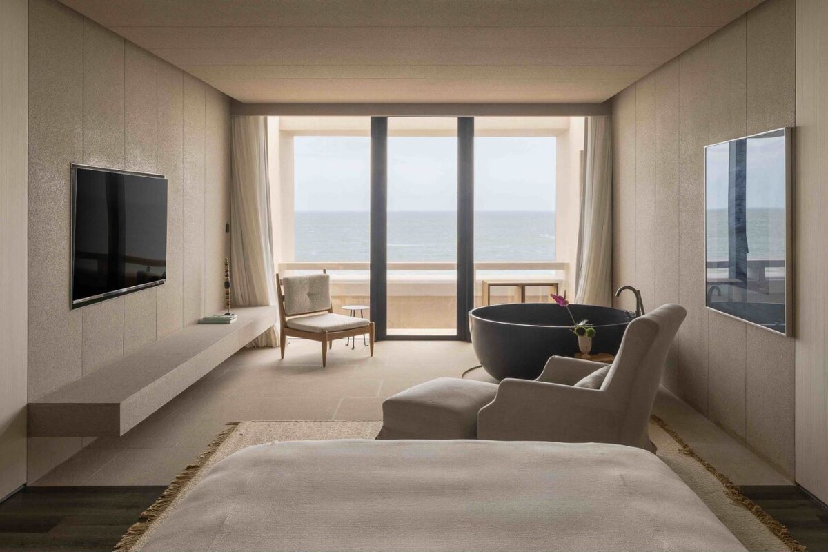 A hotel room with a bed, a TV, a chair, a table, and a view of the sea.