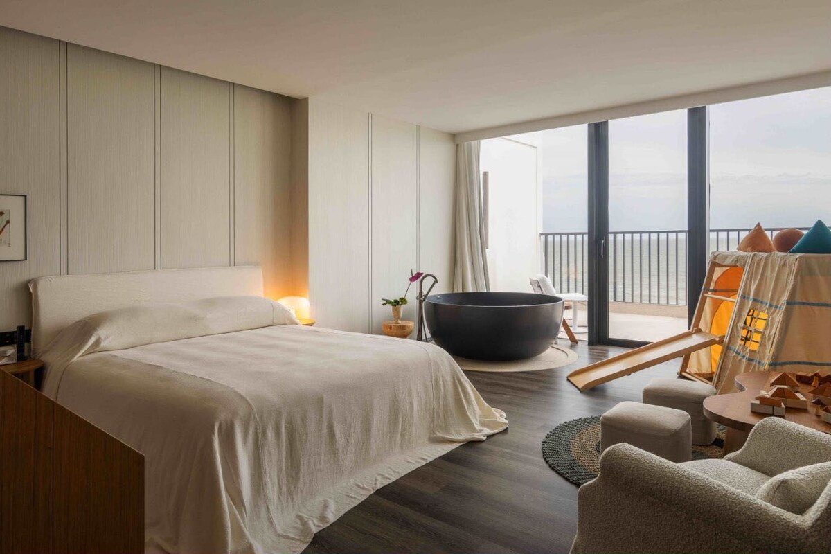 A hotel room with a large bed, a hot tub, a balcony with a view, and a seating area.