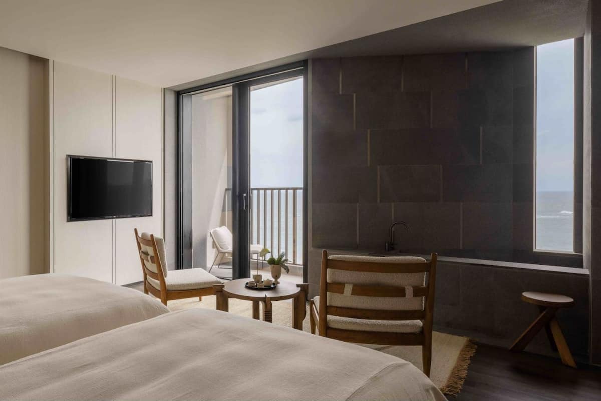 Une chambre d’hôtel avec deux lits, une télévision, un balcon avec vue, une petite table avec une chaise et un lavabo.