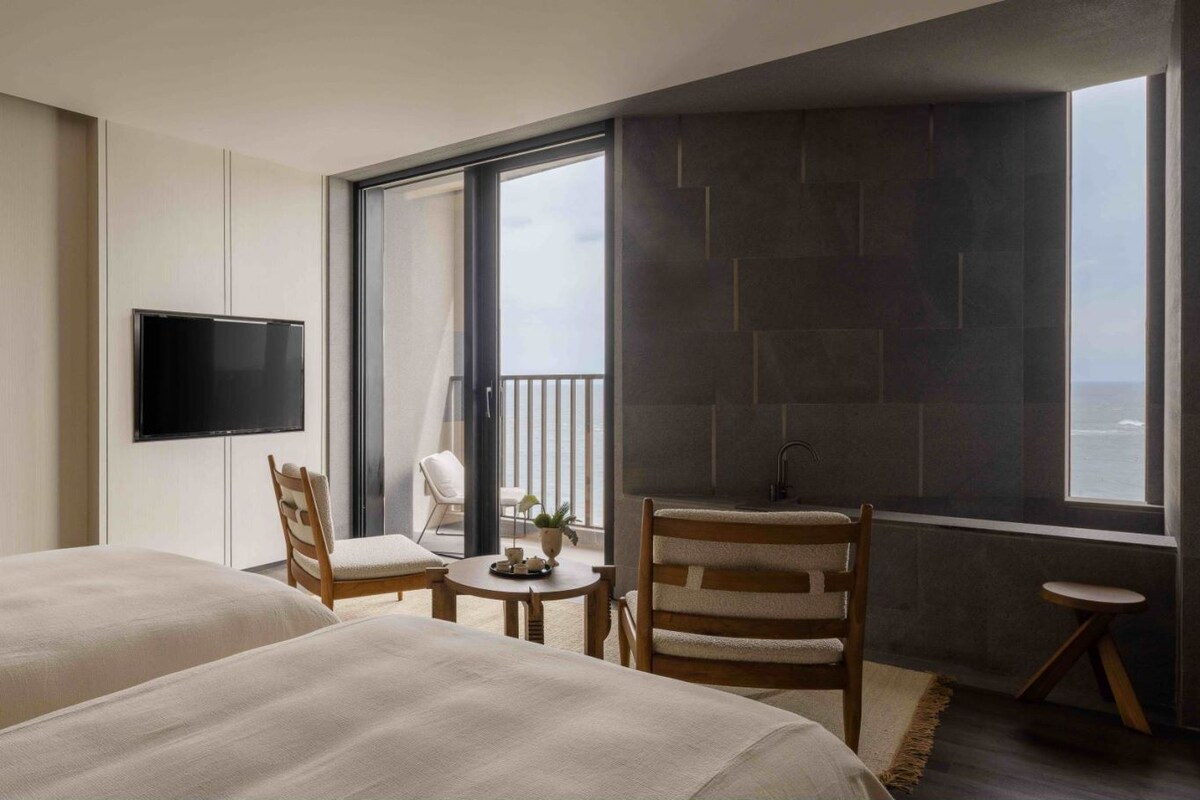 A hotel room with two beds, a TV, a balcony with a view, a small table with a chair, and a sink.