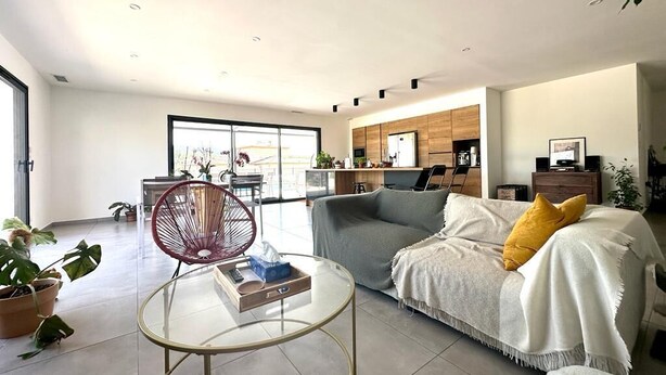 Una sala moderna con una mesa de centro de vidrio, un sofá gris con un cojín amarillo y una silla roja. Se observa una cocina al fondo con armarios de madera y una mesa de comedor.