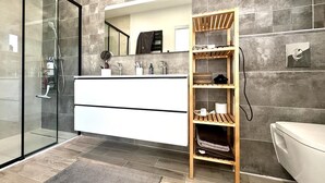 Un baño moderno con cabina de ducha de vidrio, tocador blanco con espejo y estante de madera.