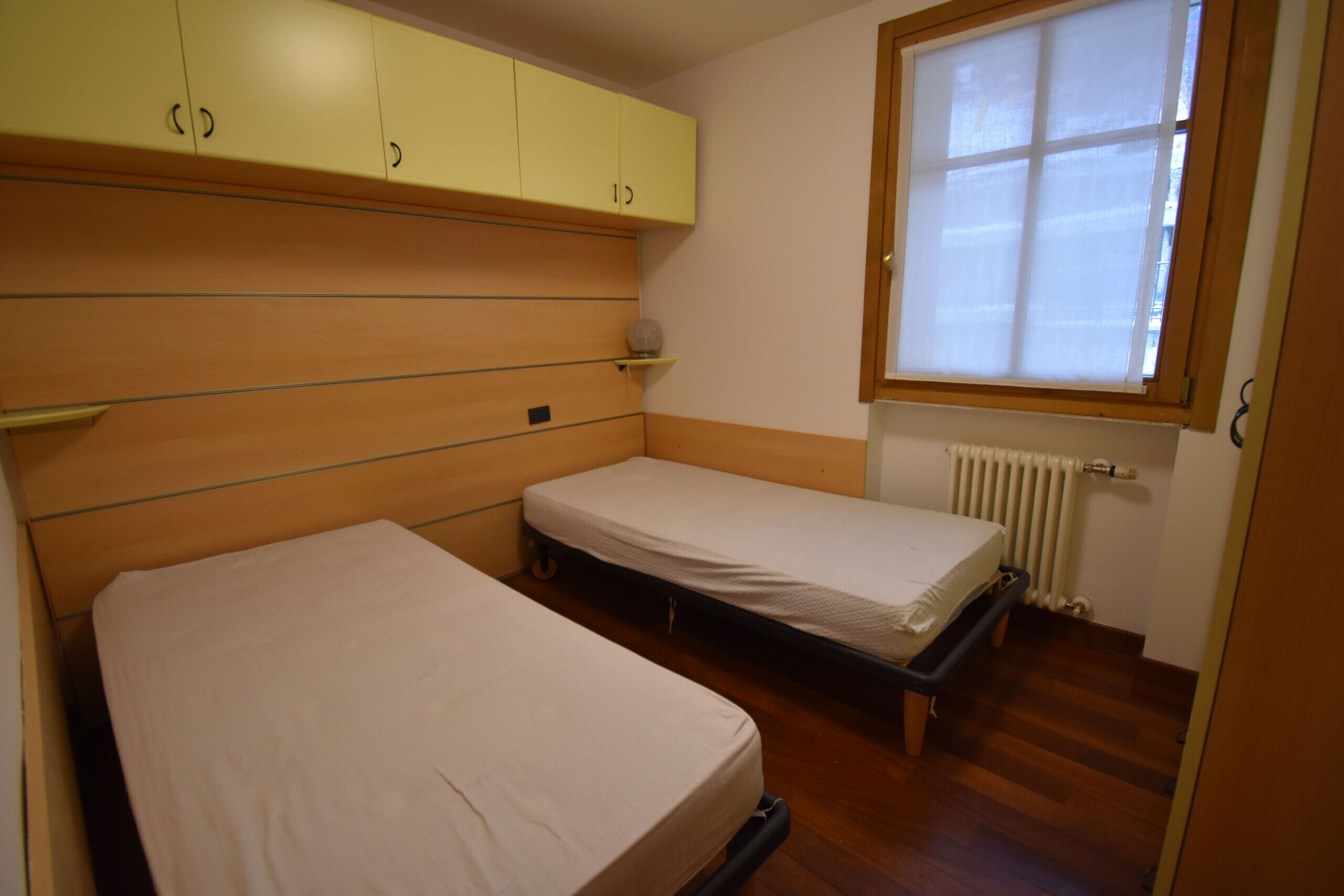 A small bedroom with two single beds, wooden cabinets, a window, and a radiator.