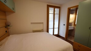 Een slaapkamer met een bed, een radiator, een kledingkast en een badkamer, zichtbaar door een deur.