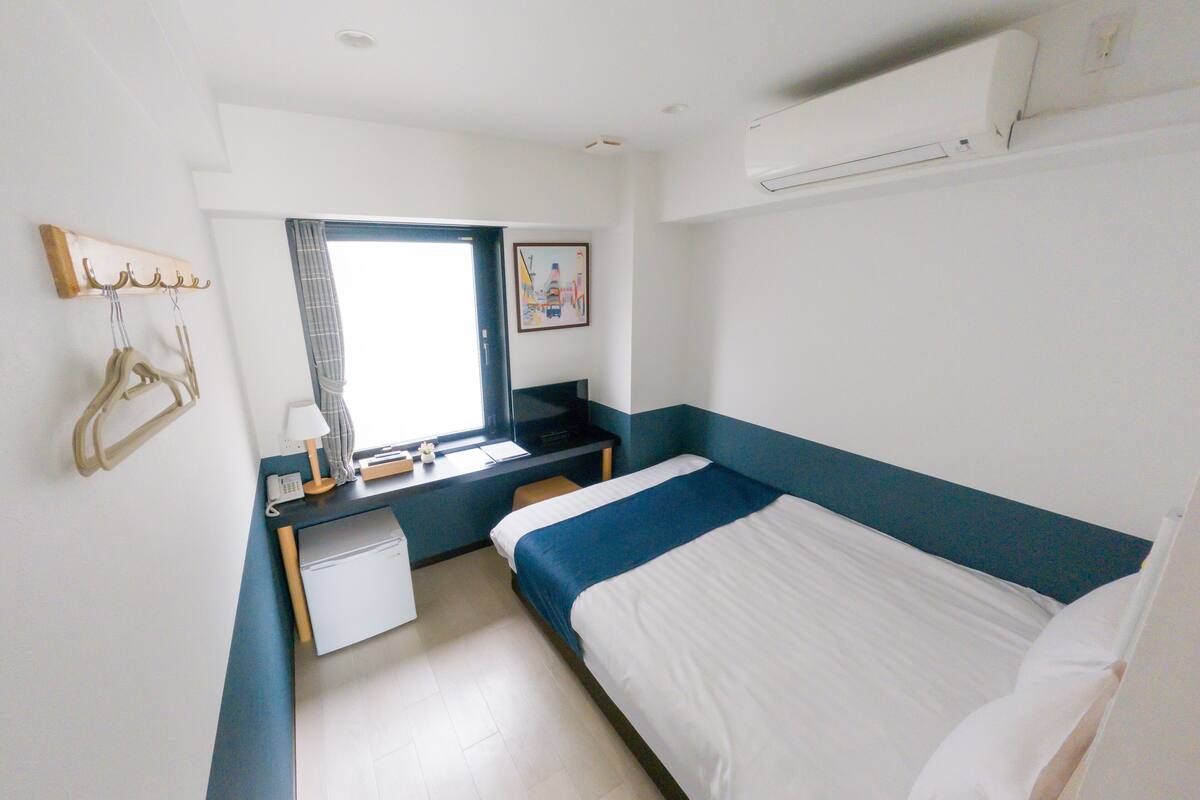 A small, clean room with a bed, a desk, a chair, a small table, and a window.