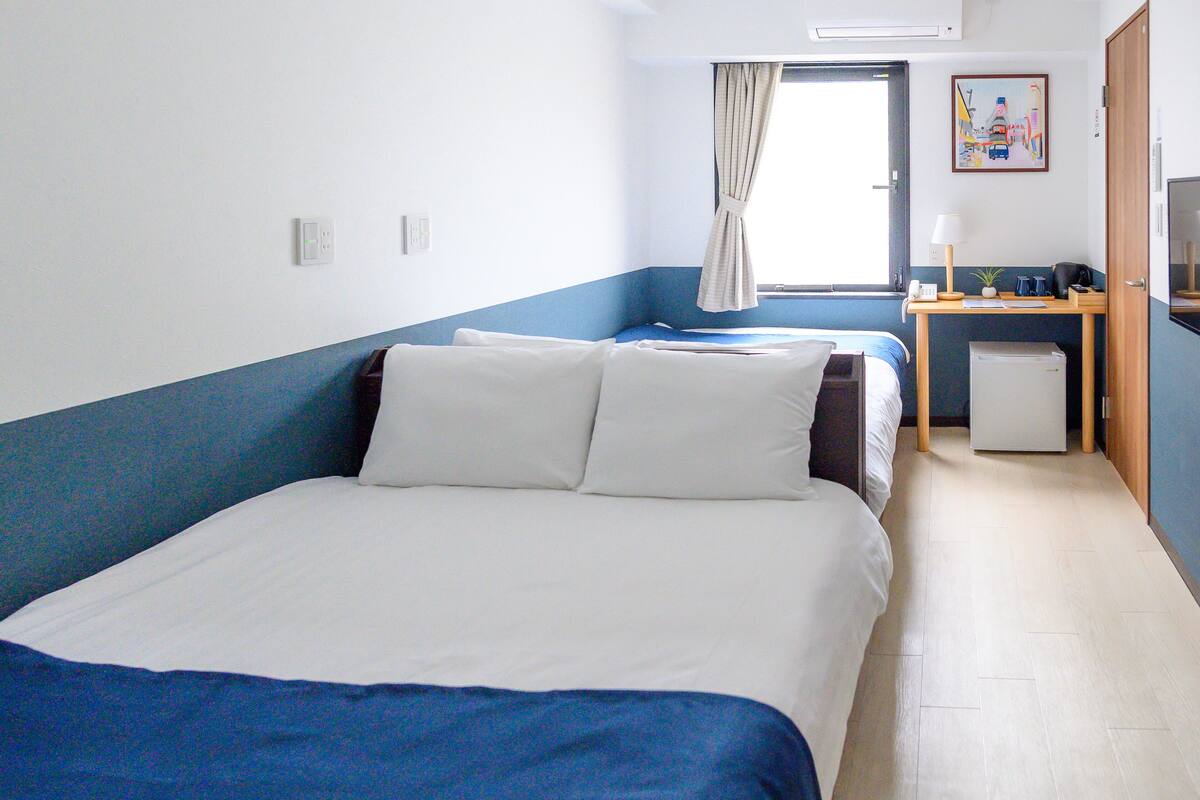 A hotel room with a large bed, a desk with a chair, a small refrigerator, and a window with curtains.