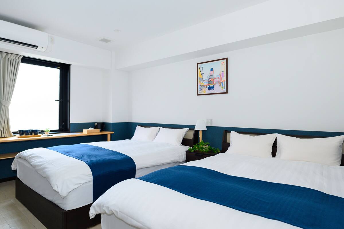 A hotel room with two beds, a window with curtains, a painting on the wall, and an air conditioning unit.