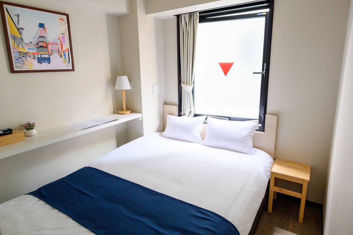 A hotel room with a bed, a bedside table, a lamp, a small plant, and a window with curtains.