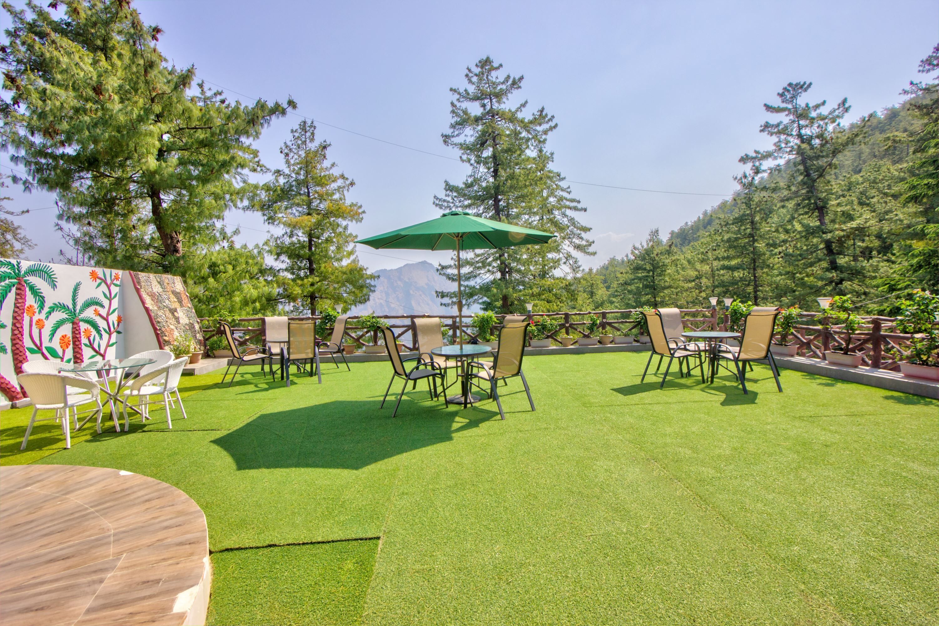 An outdoor seating area with artificial grass, tables, and chairs, surrounded by trees and a mountain view.