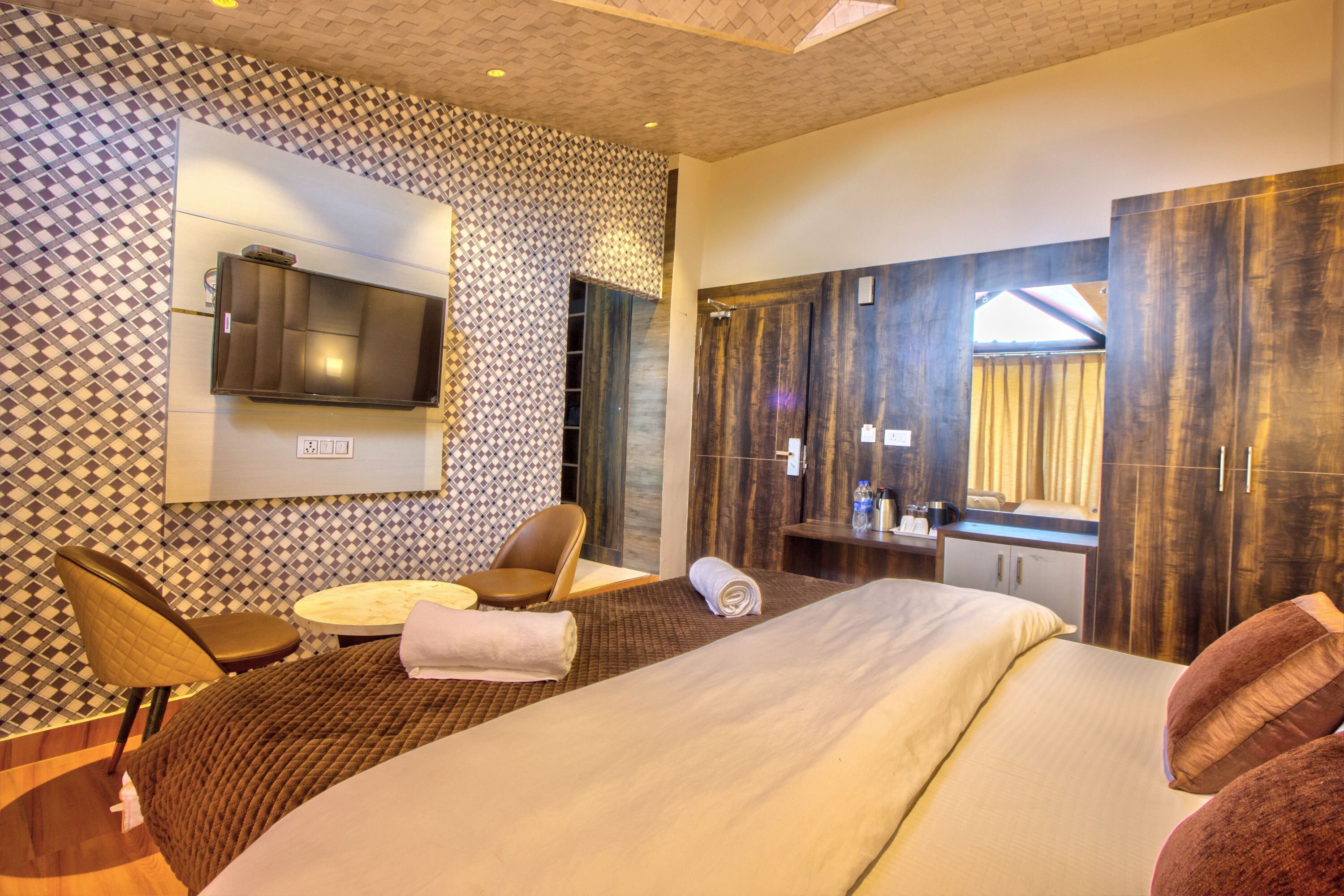 A hotel room with a bed, a TV mounted on the wall, a seating area with chairs, a small table, and a bathroom with a mirror and curtains.