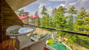 Un balcon agrémenté de meubles en osier, d’une table en verre et offrant une vue sur un paysage verdoyant, avec des arbres et un bâtiment doté d’un toit rouge.