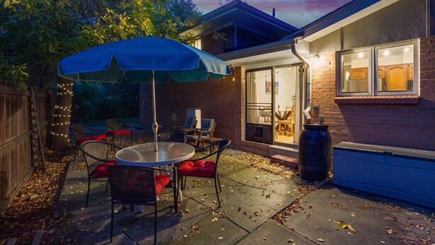 An outdoor patio with a table and chairs, a blue umbrella, and a brick house in the background.