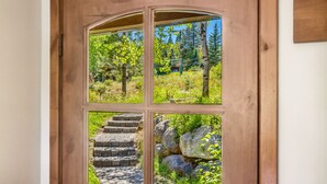 Una puerta de madera con un panel de vidrio, mostrando una escalera de piedra y un espacio natural exterior con árboles y rocas.