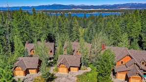 Un conjunto de casas tipo cabaña de troncos con garajes adjuntos, rodeado de un bosque denso y un lago con montañas al fondo.