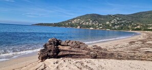 Ein Strand mit einer großen Felsformation im Vordergrund und einer hügeligen Landschaft im Hintergrund.