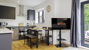 A modern kitchen with dark cabinets, a built-in oven, and a dining area with a table and chairs. A flat-screen TV is mounted on the wall, and a clock is visible.
