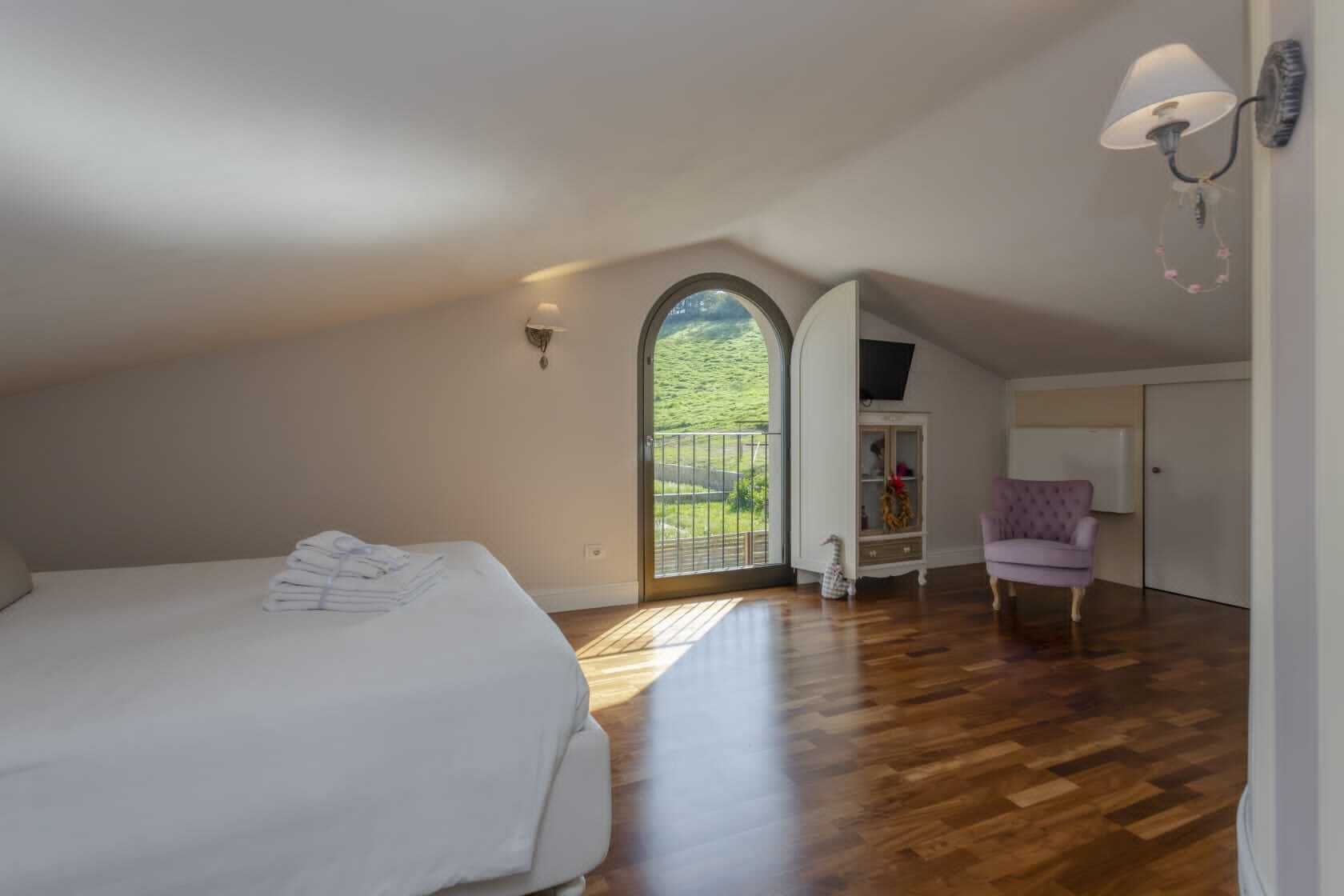 A bedroom with a large bed, a wardrobe, a chair, and a view of a green landscape through a window.