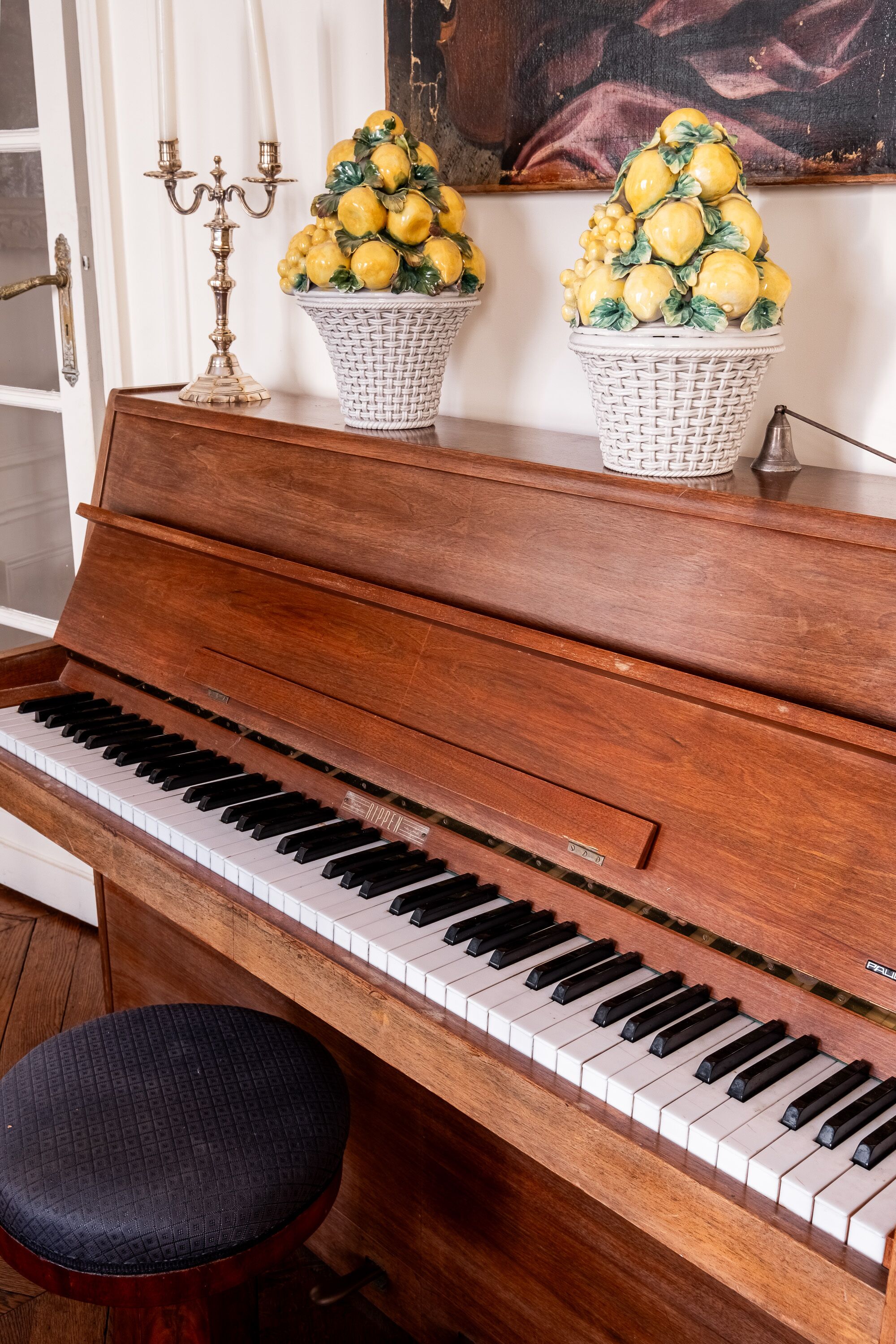 A grand piano with a black cushioned bench, two wicker baskets with yellow lemons, and a candelabra on a wooden table.