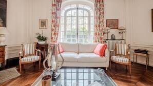 Et værelse med en hvid sofa, to stribede lænestole, et glasbord og et stort vindue med blomstrede gardiner.