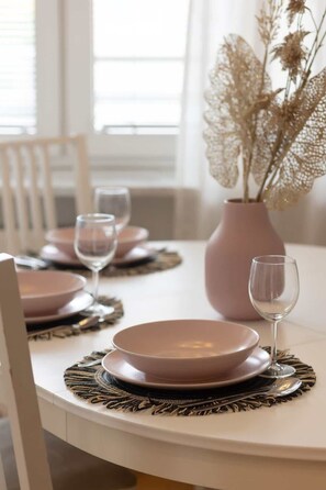 Una mesa de comedor dispuesta con platos rosas, cubiertos y copas de vino, junto con un jarrón con flores secas.