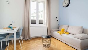 A modern living room with a sofa, a dining table with chairs, a round side table, a window with curtains, and a wall clock. - Modern Studio in Kazimierz | 1 km from the Wawel Castle (Kraków)