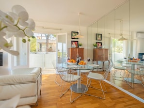 A modern dining area with a glass table, metal chairs, and a wooden cabinet with artwork. - Apartamento Bonaire · Your beachfront apartment in Alcúdia. (Alcúdia)