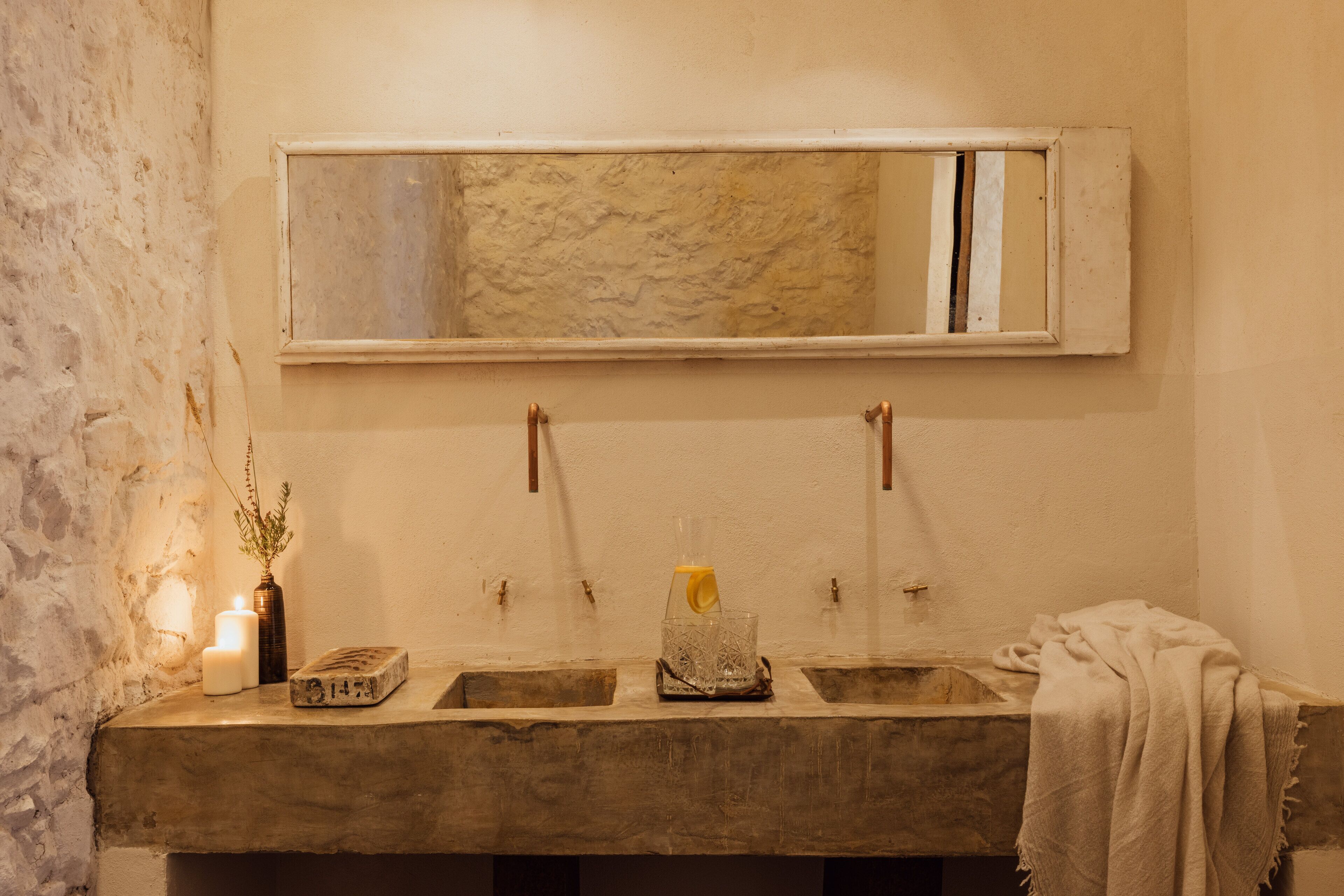 A bathroom with a large mirror, two sinks, a candle, a vase with a plant, and a towel.