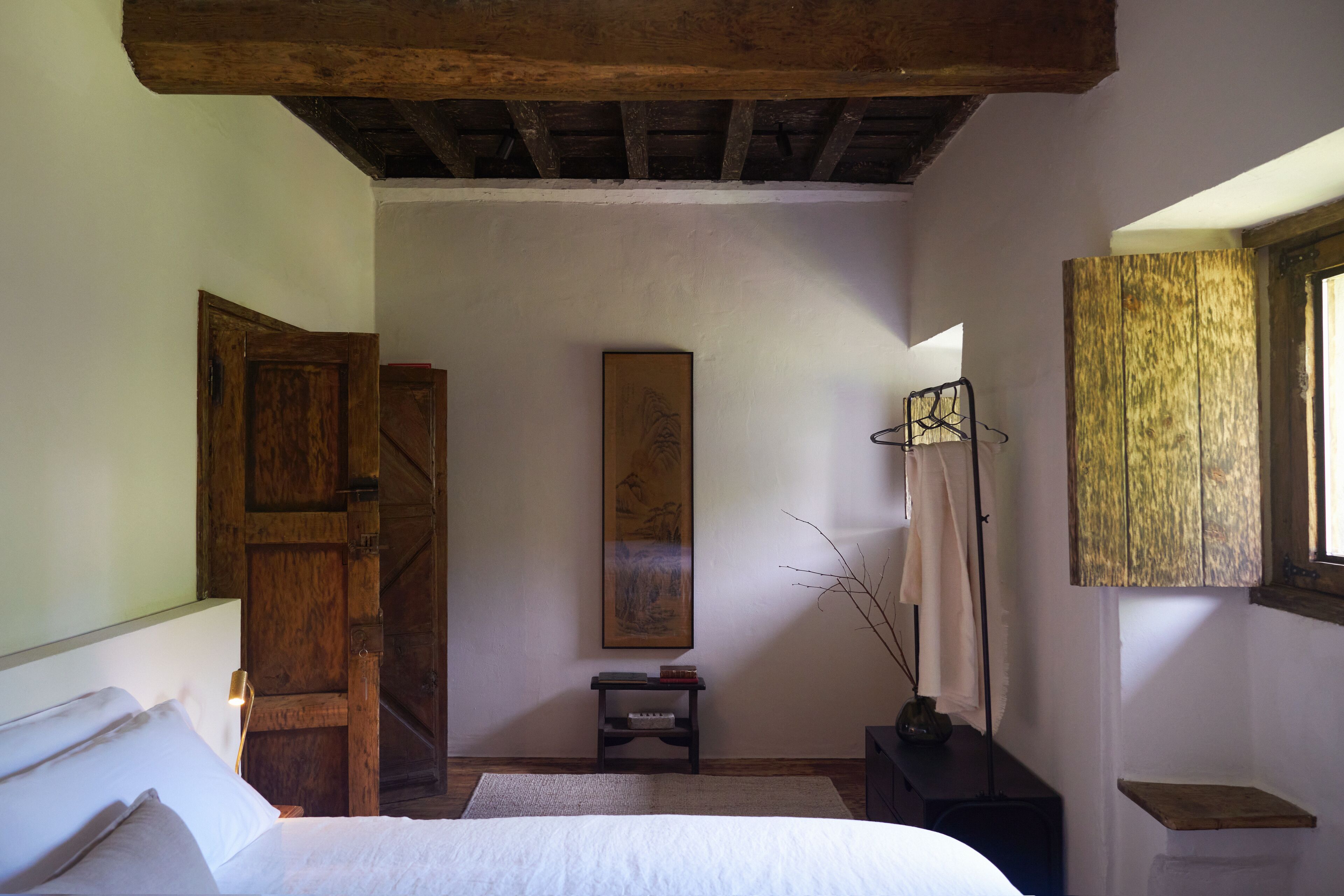 A bedroom with a wooden door, a bed, a small table with a vase, and a painting on the wall.
