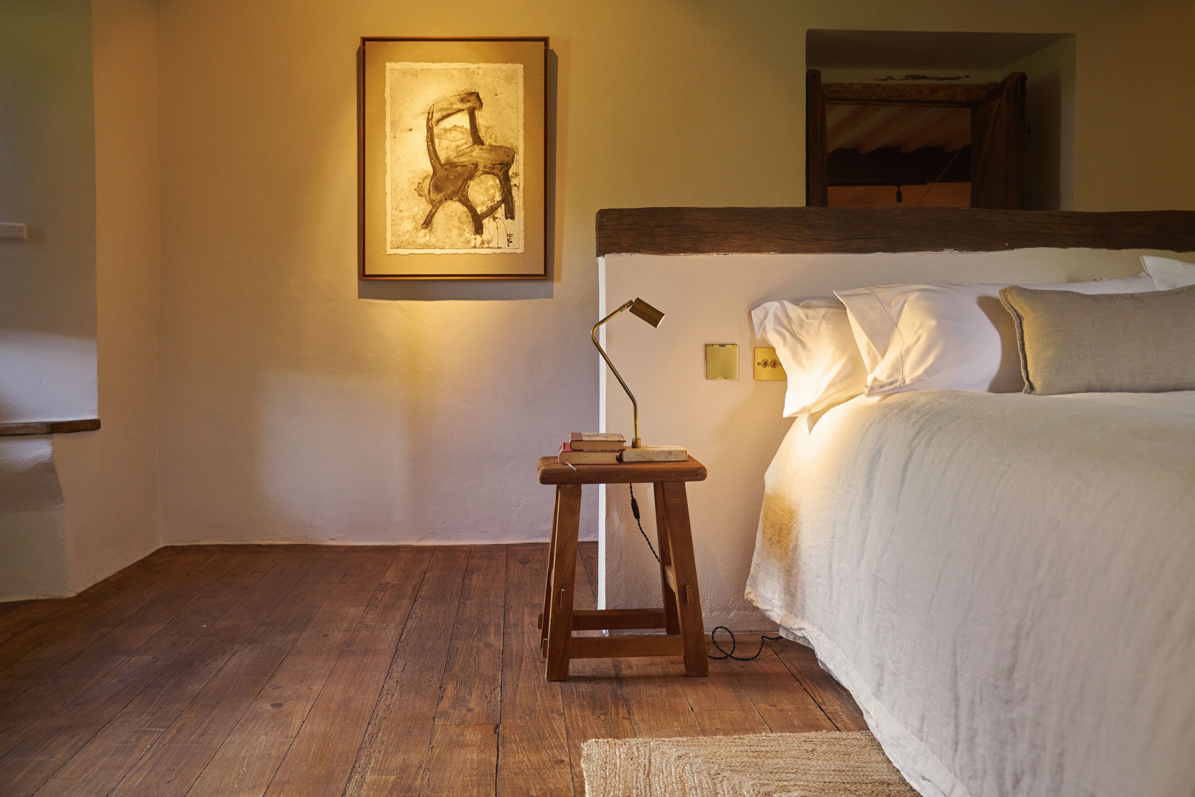 Een slaapkamer met een houten vloer, een bed met witte beddengoed, een klein houten krukje met boeken, een ingelijst schilderij aan de muur, en een bedlampje.