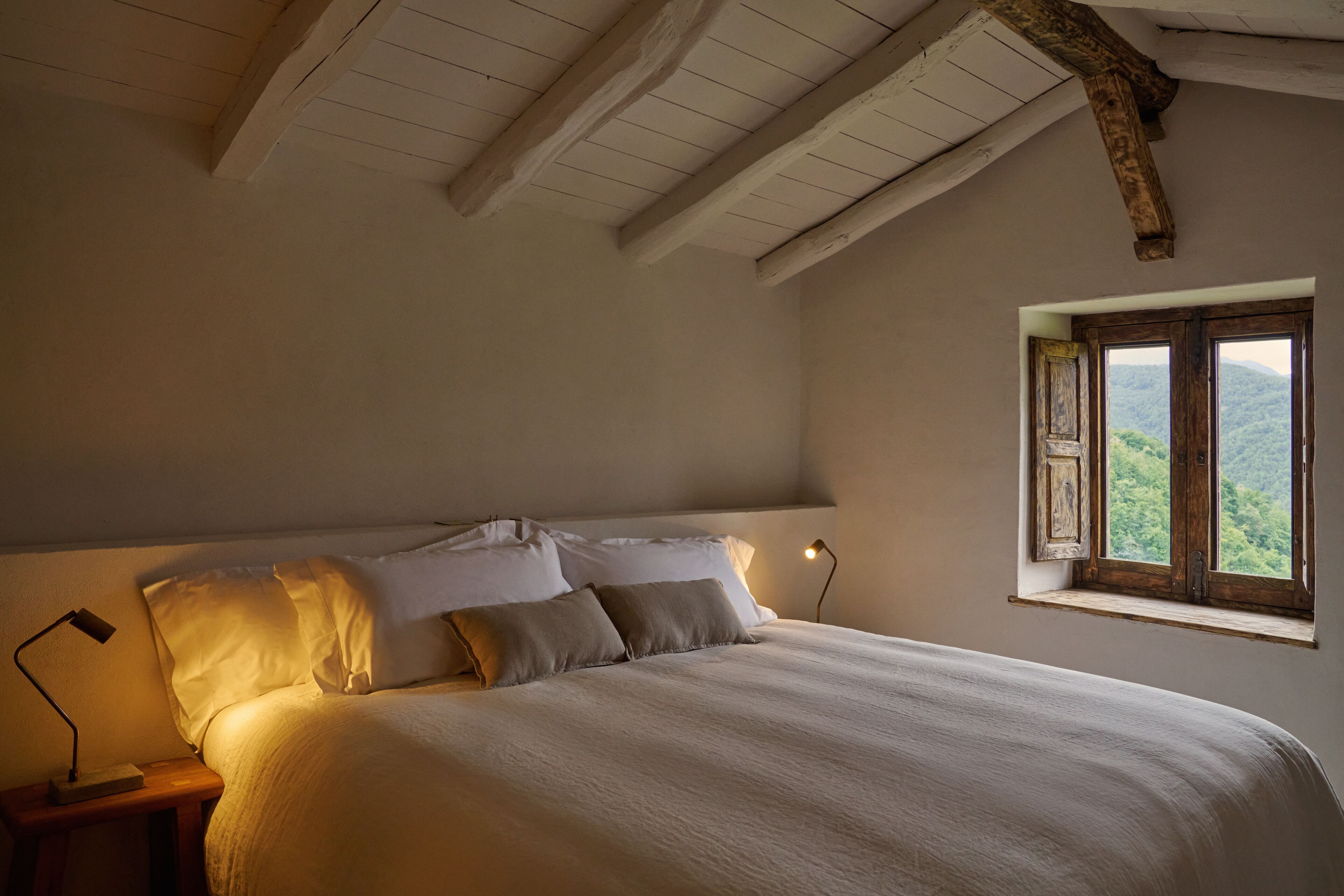A bedroom with a large bed, a nightstand with a lamp, and a window with a view of greenery.