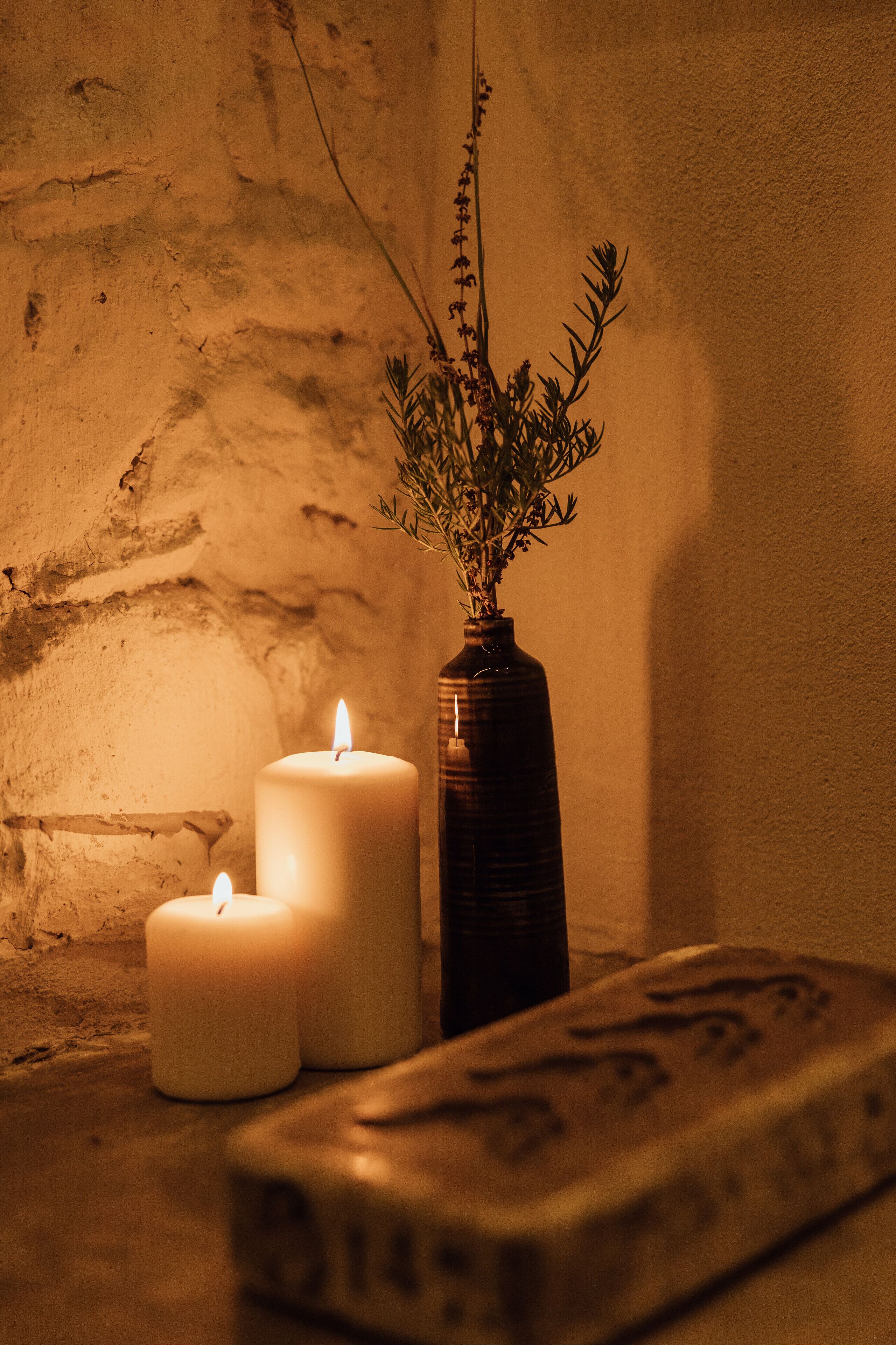 Escena iluminada por velas con un florero de plantas secas y dos velas encendidas sobre una pared con textura.